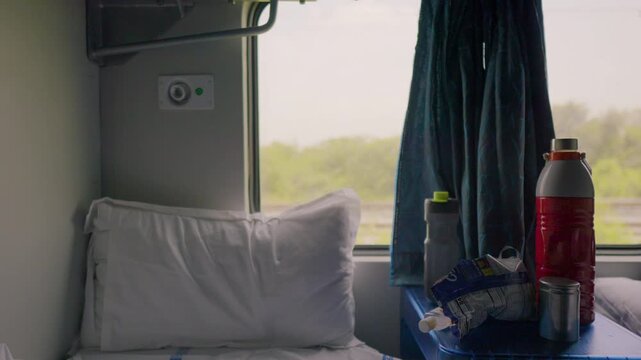 An authentic interior view of travel snacks, water bottles, and a thermos on a cabin side table, framed against a white bedroll pillow and passing green scenery through the train window.