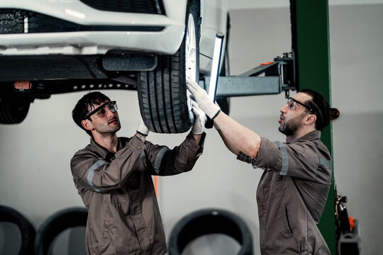 Two Mechanics Working Together Underneath Car Suspended on Lift.  Auto Repair Team Collaborating on Brake and Chassis Maintenance in Workshop