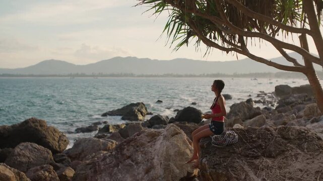 Young asian woman with long braids sits on rocky seashore holding a yellow cassette player beneath a coastal seashore screw pine tree, golden hour dusk sunset glow