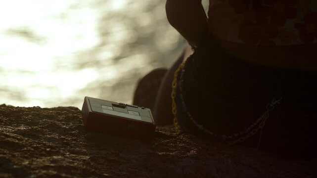 Red portable cassette player rests on a coastal rock beside a young woman at sunset, closeup abstract as woman sits on cliffside