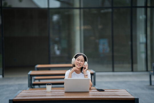 A smiling young woman wearing headphones works remotely on her laptop, studying and communicating online while enjoying music&mdash;an image that reflects the use of technology, work efficiency,