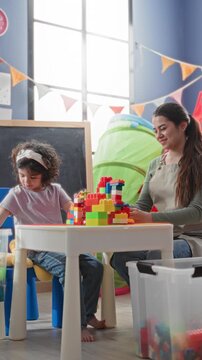 Woman supervises girl stacking colorful plastic blocks on a small table in a classroom building with chalkboard and playtent visible; learning joy.