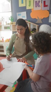 Teacher writing on paper with green pencil while child cuts paper at table in classroom; nurturing concentration.