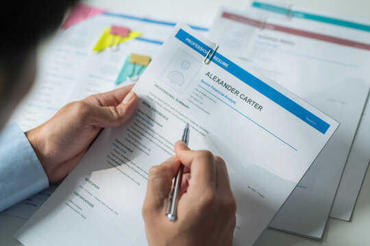 Close-up of worker holding pen writing on report and inspecting resume documents at office desk.