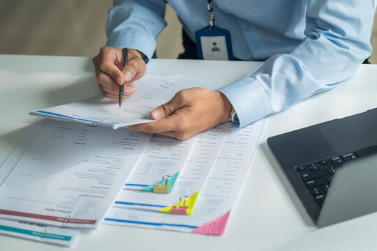 Close-up of worker holding pen writing on report and inspecting resume documents at office desk.