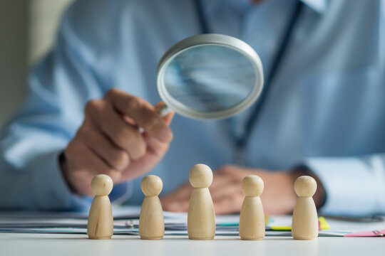 Close-up of wooden dolls on desk with worker holding magnifying glass to inspect blurred in background.