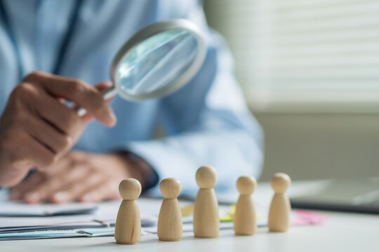 Close-up of wooden dolls on desk with worker holding magnifying glass to inspect blurred in background.