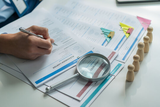 Close-up of magnifying glass and wooden dolls on resume documents with worker holding pen at desk.