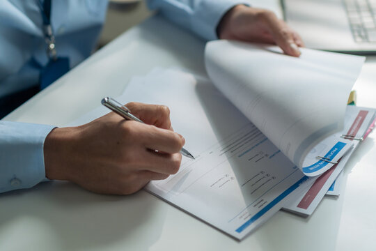 Close-up of worker holding pen and flipping paper to check report documents at desk.