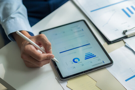 Close-up of worker holding stylus pen writing on digital tablet screen to check financial report at desk.