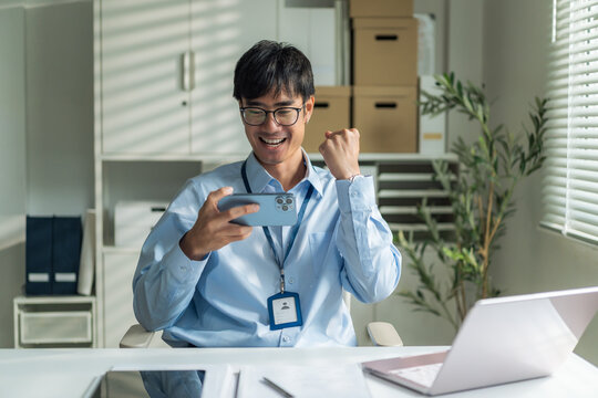 Office worker wearing glasses using smartphone horizontally and gesturing fist pump celebrating success at desk.