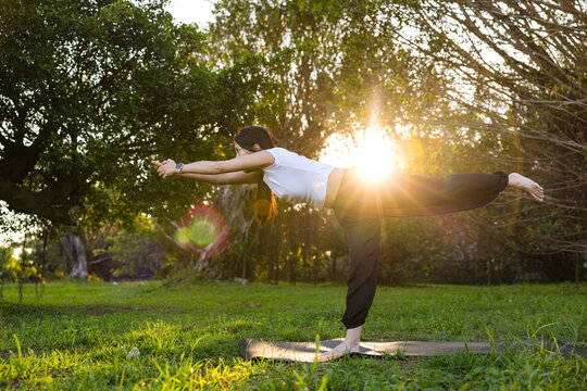Woman balancing in a warrior three yoga pose on a lawn during golden hour