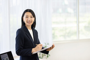 Obraz premium Young Japanese businesswoman holding a clipboard and smiling at the camera by a bright window