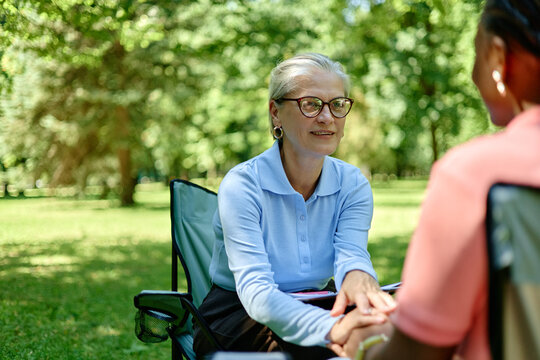 Senior Caucasian female therapist holding hands of Black female patient during outdoor psychotherapy session in park, promoting mental health support and ecotherapy