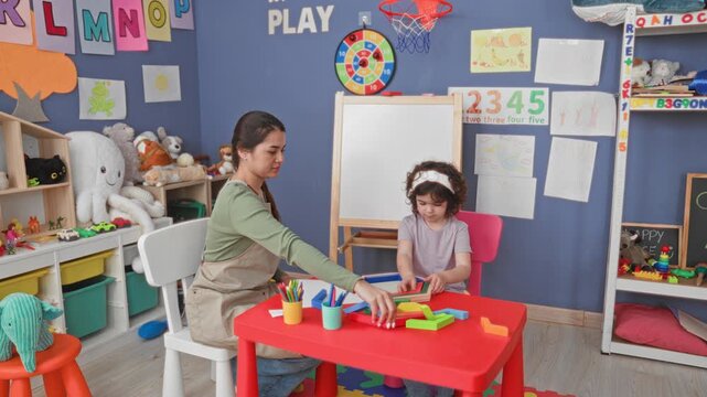 Woman guiding girl stacking colorful wooden blocks on a red table in a building; concentration learning.