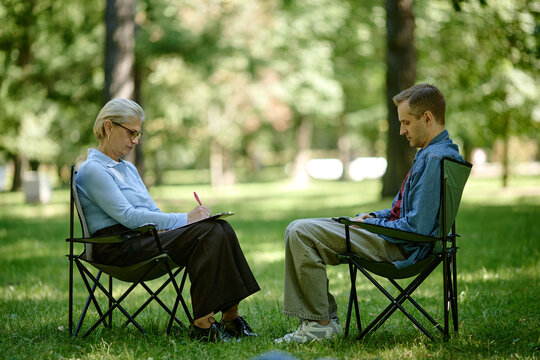 Senior Caucasian female therapist taking notes during outdoor psychotherapy session with Caucasian man in park, representing mental health support in nature