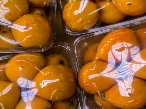 High-angle close-up of ripe yellow loquats (Eriobotrya japonica) neatly arranged and wrapped in transparent plastic film for sale inside a grocery store or traditional supermarket stall in Brazil.