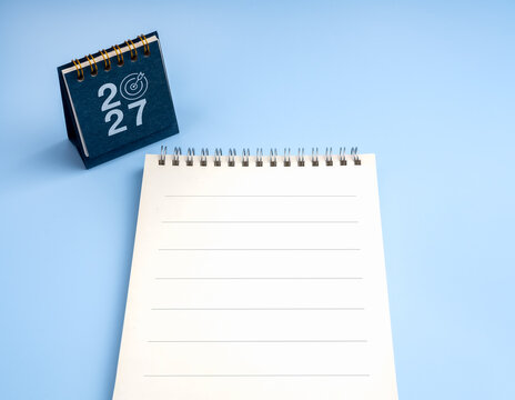 Blank lined notebook beside 2027 desk calendar with target symbol on blue background, representing annual planning, goal setting, scheduling, to do list, resolutions and organized preparation concept.