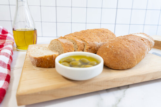 Fresh bread rests on a wooden board. Small bowl holds green olives and olive oil. Red-and-white checked cloth adds rustic charm.