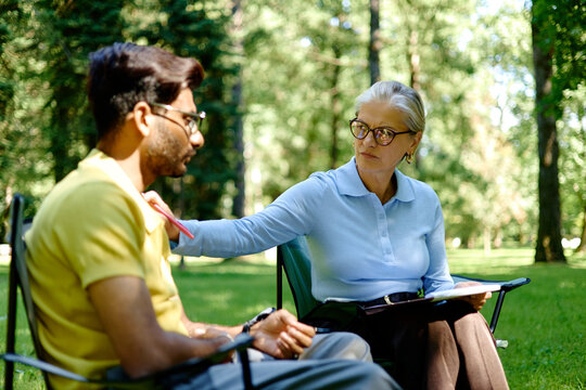 Senior Caucasian female therapist comforting young adult Indian male patient during outdoor psychotherapy session in park, perfect for mental health and counseling services promotion