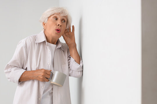 Hearing loss and senior health, elderly woman eavesdropping through a wall with a coffee mug in hand to listen to secrets or private conversation