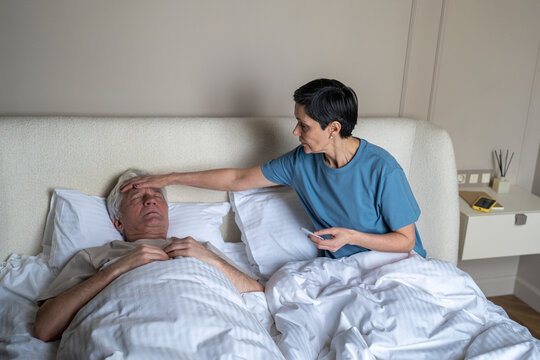Anxious mature wife taking temperature of ill feverish husband in bed with thermometer. Health monitoring, caregiving spousal concern, home sickness, family care, medical attention, intimate support
