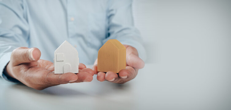 Business professional holding two wooden house models comparing real estate options, concept of property choice, home buying decision, mortgage comparison, and housing investment.