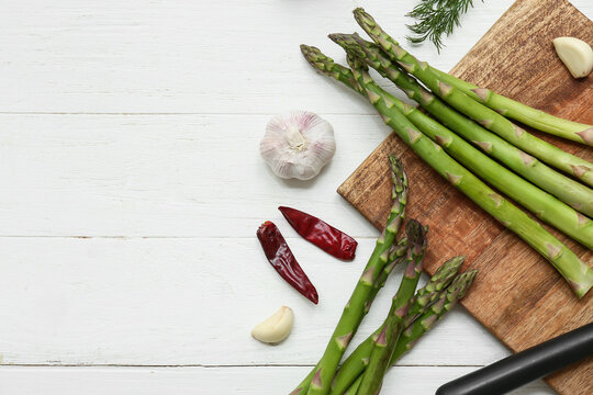 Cutting board with fresh green asparagus and different spices on white wooden background