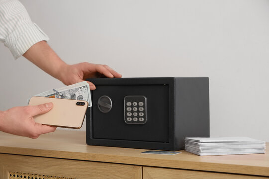 Man putting money with mobile phone in safe on shelf near light wall, closeup