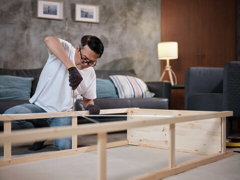 Asian mature man concentrates on self-assembling wooden furniture with DIY tools, tightening strength with a screwdriver, home improvement, hobby carpentry workshop, expert handyman, active lifestyle.