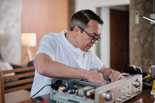 Mature Asian man disassembling and examining audio equipment, checking circuit board for repair, home electrical appliance DIY fix and maintenance, electronics component hobby, handyman workshop.