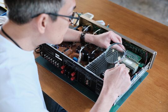 Close-up, man's hand disassembling audio equipment, checking circuit board for repair with a magnifying glass, appliance DIY fix and maintenance, electronics component hobby, handyman workshop.