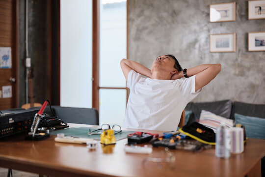 Mature Asian man concentrates on repairing home electrical appliance, disassembling and soldering circuit board, tired from precision detail focusing, taking a break, resting from his workshop hobby.