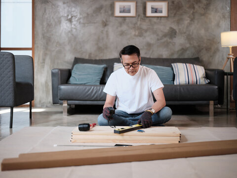 Asian mature man sitting and preparing flat-pack furniture parts and DIY equipment for self-assembly, home improvement, wooden craft installation, hobby workshop, carpentry handyman active lifestyle.