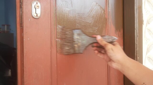 A man's hand is applying brown wood coating paint to the surface of a wooden panel door.