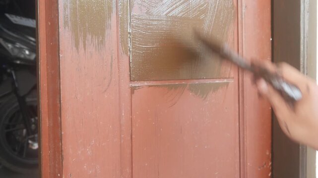 A man's hand is applying brown wood coating paint to the surface of a wooden panel door.