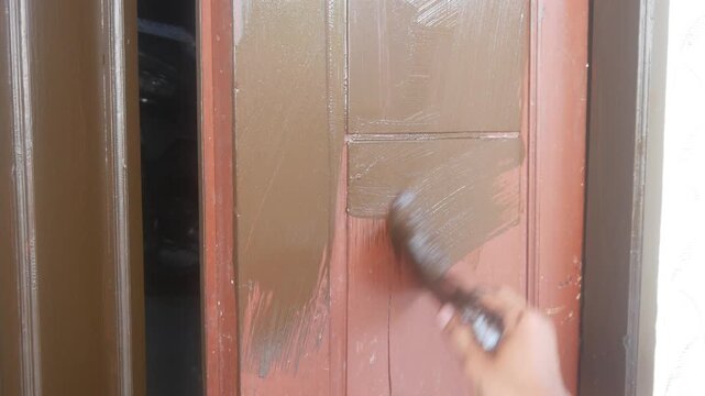 A man's hand is applying brown wood coating paint to the surface of a wooden panel door.