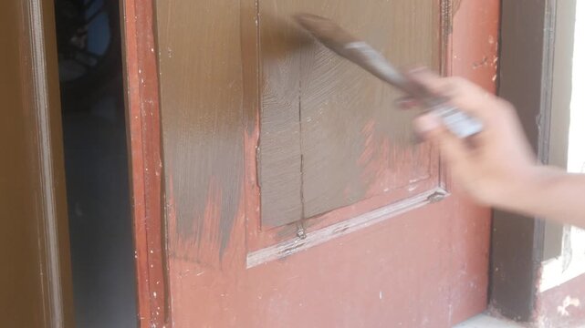 A man's hand is applying brown wood coating paint to the surface of a wooden panel door.