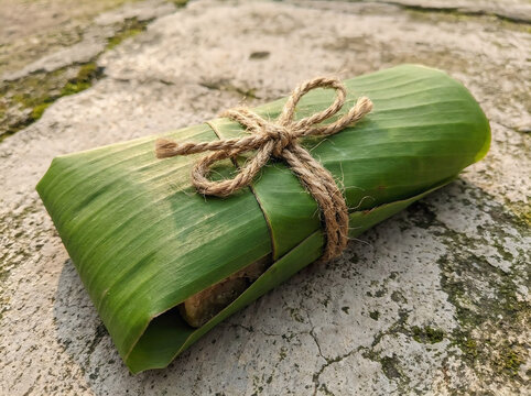 Traditional sticky rice snack wrapped in a vibrant fresh green banana leaf with a rustic jute string bow tied neatly atop a weathered stone floor surface.