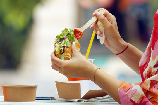 Elegant woman with manicured hands and colorful floral outfit drizzling red sauce onto a loaded street food sandwich, savoring an unhurried moment at Karlin food festival in Prague, Czech Republic. 