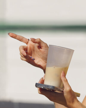 A woman gestures with one hand while holding a chilled iced coffee and her phone in the other, a candid on-the-go moment at the sunny Naplavka farmers market in Prague.