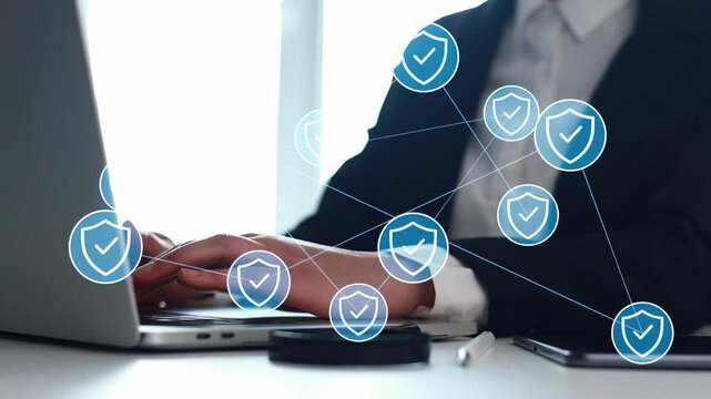 A person works on a laptop in an office while digital icons of shields with check marks float around. The scene shows a focus on cybersecurity and technology use.