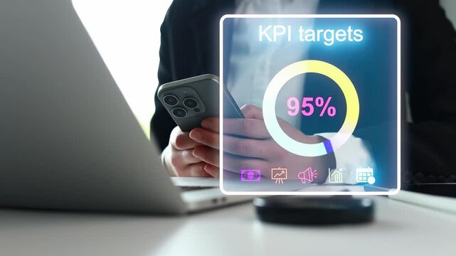 A person looks at their smartphone while seated at a desk. The phone displays a KPI target of 95 percent. There is a laptop and a round device on the desk.