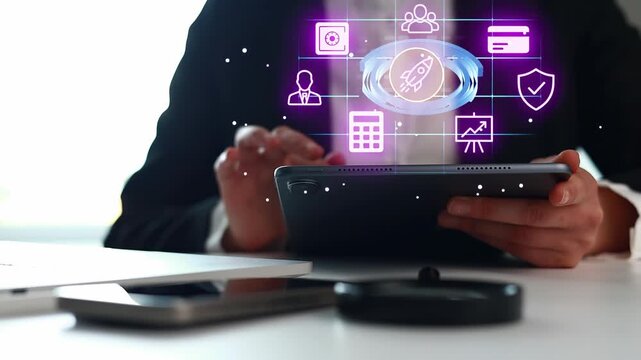 A person holds a tablet and looks at digital icons floating above the screen. The workspace has a clean setup with other devices like phones and notebooks nearby.