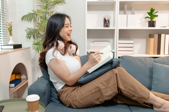 Young Asian woman wearing wireless headphone while reading a book on cozy living room sofa. Peaceful home lifestyle representing self learning, audiobook listening, relaxation and personal development