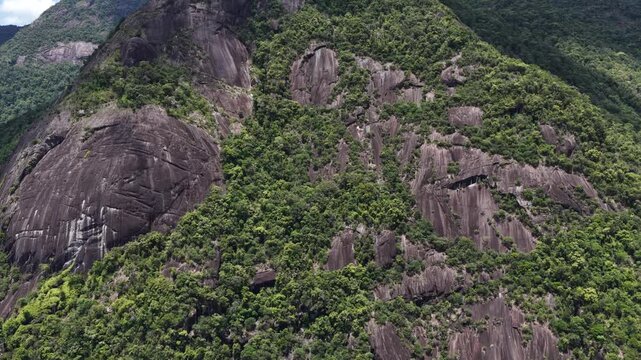 High Aerial Drone View of Yang Hanh Mountain in Dak Lak, Vietnam, Revealing the Entire Rugged Peak and a Massive Monolithic Rock Formation on the Left Beneath Moving Patches of Sunlight and Cloud