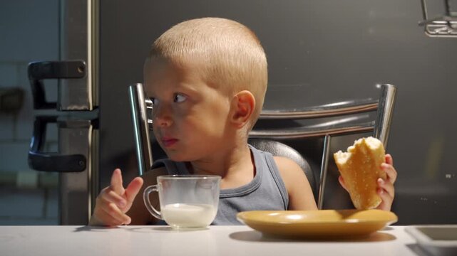 Young toddler seated on metal chair at kitchen counter holds bread roll in right hand and points with left hand while small glass cup of milk rests nearby, creating warm domestic morning scene with