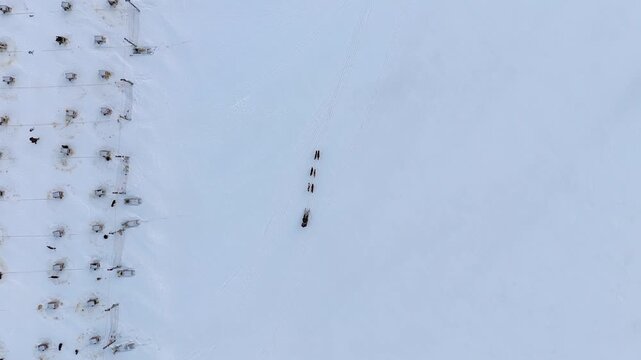 Aerial drone footage descending slowly over a snow covered field in Svalbard, showing a sled dog team pulling a musher across the open snow near the kennel yard