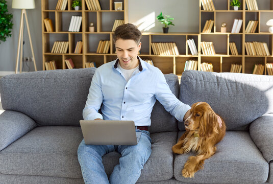 Man with dog doing remote work at home. On a sofa with laptop, smiling in a bright room. Cozy lifestyle shows companionship and productivity for online tasks. Remote work with pet at home.