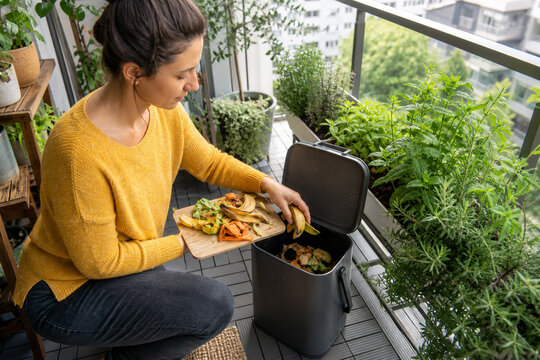 Apartment Resident Adding Fruit and Vegetable Kitchen Scraps to a Compact Sealed Balcony Compost Bin Beside Healthy Herb Planters